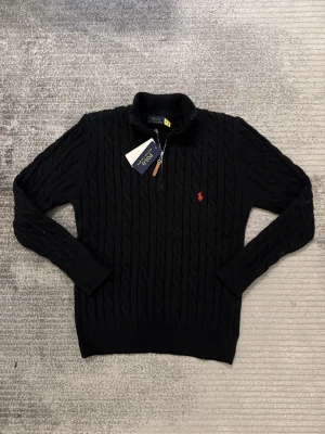 Svart stickad tröja från Polo Ralph Lauren - Svart kabelstickad tröja från Polo Ralph Lauren med halv dragkedja och klassisk röd logga broderad på bröstet. Tröjan har hög krage och är långärmad, perfekt för kyligare dagar. Materialet är mjukt och värmande ull.