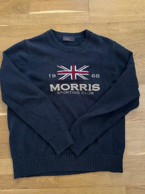 Mörkblå stickad tröja Morris L - Säljer en mörkblå stickad tröja från Morris Sporting Club i storlek L. Tröjan har ett broderat Union Jack-flagga och texten 'Morris Sporting Club 1968' på bröstet. Klassisk rund halsringning och ribbade muddar vid ärmslut och nederkant. Sitter inte jätte stort 