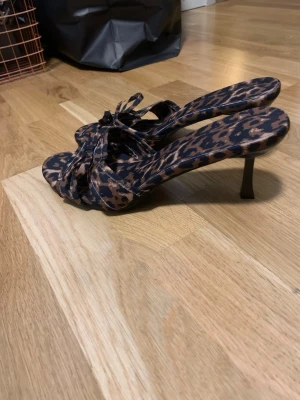 Leopardmönstrade sandaler med klack - Säljer ett par sandaler i storlek 39 med leopardmönster och öppen tå. Skorna har smal hög klack och en söt rosettdetalj framtill. Materialet är syntet och de har en cool brun och svart färgkombination. Perfekta för dig som vill sticka ut lite extra.