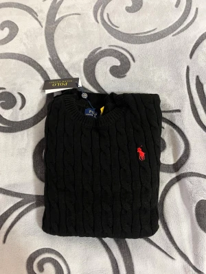 Svart kabelstickad tröja Polo Ralph Lauren - Kabelstickad svart tröja från Polo Ralph Lauren med klassisk röd logga broderad på bröstet. Tröjan är i 100% bomull, har rund halsringning och långa ärmar. Perfekt för dig som gillar stilrena och tidlösa plagg.