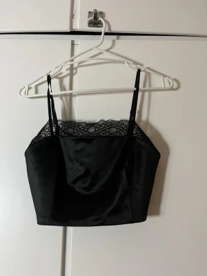 Svart satin bandeau topp med spets - Säljer en svart bandeau topp i satin med tunna axelband och snygg spetskant upptill. Toppen har en croppad passform och smockad rygg för extra komfort. Perfekt för dig som vill ha en stilren och trendig look.