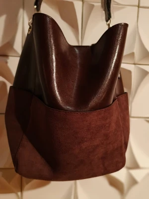 Helt ny Åhléns axelväska i vinrött! - Elegant och fin handväska från Åhléns i vinrött, tillverkad i skinnimitation och mockaliknande material.  Rymlig och mjuk modell - perfekt både till vardags och för en mer elegant stil.  Justerbar axelrem och magnetstängning.  En längre och smalare axelrem tillkommer även i väskan som man kan byta ut!  Skick: Ny med tagg  Ordinarie pris: 599 SEK  Storlek: ca 35 x 30 x 14 cm