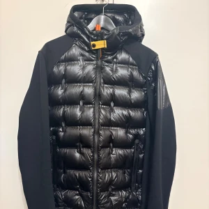Svart dunjacka från Parajumpers - Snygg svart dunjacka från Parajumpers med glansig quiltad framsida och huva. Jackan har svarta ärmar i kontrastmaterial, dragkedja framtill och en gul detalj vid halsen. Perfekt för kalla dagar när du vill ha både stil och värme. Extra knappar Medföljer!!, köpt för 6799kr