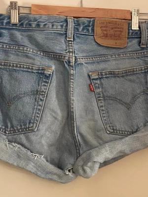 Levi's ljusblå denimshorts - Säljer ett par klassiska ljusblå denimshorts från Levi's med upprullade benslut och femficksdesign. Shortsen har hög midja, synliga slitningar och den ikoniska läderpatchen bak. Perfekta för varma dagar och en avslappnad utseende.