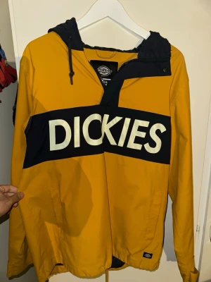 Gul och marinblå vindjacka från Dickies - Säljer en snygg gul och marinblå vindjacka från Dickies i storlek small men passar M också. Jackan har stor logga över bröstet, huva med snörning och dragkedja upptill. Praktiska fickor på sidorna och justerbar nederkant med snöre. Perfekt för dig som vill sticka ut.