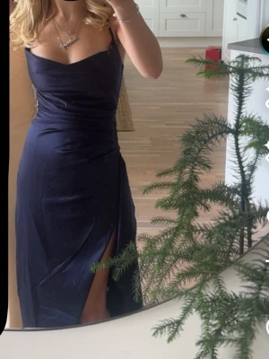 Marinblå balklänning med slits - Säljer en marinblå balklänning med smala axelband och snygg slits framtill. Klänningen har en elegant, figurnära passform och ett mjukt, glansigt tyg som ger en lyxig känsla. Perfekt för bal