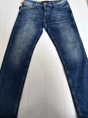 Blå jeans från Jack & Jones - Säljer ett par klassiska blå jeans från Jack & Jones med snygg tvätt och subtila slitningar. Modellen har fem fickor, guldfärgad knapp och raka ben. Jeansen är tillverkade i bomull och har en normal passform som funkar till det mesta. Lappen kvar aldrig använda