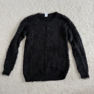 Svart fluffig stickad tröja Vero Moda - Mysig svart stickad tröja från Vero Moda i storlek XS. Tröjan har en fluffig och lite lurvig textur som gör den extra gosig. Långärmad modell med rund halsringning och ribbade muddar. Perfekt för kyliga dagar!