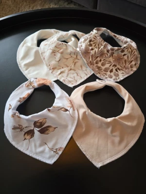 Beige och catalinablommiga dregglisar  - Fem mjuka dregglisar i triangelform, i olika beige och vita toner. Några har blommigt mönster i brunt och beige, andra är enfärgade. Dregglisarna har tryckknappar och är sydda i bomullsmix med fleecebaksida perfekta för en stilren look. Halsomkrets ca 33-34 cm. 