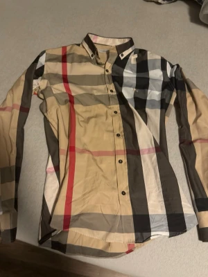 Rutig skjorta från Burberry London L - Säljer en klassisk rutig skjorta från Burberry London i storlek L. Skjortan har beige, svart, vit och röd färg i det ikoniska Burberry-mönstret. Tillverkad i mjuk bomull med knappar framtill och lång ärm. Perfekt för dig som vill ha en tidlös och snygg look. Det står xl men jag sydde den så den passar L priset går att sänka det är bakdelen av skjortan som är sydd och både armen men den passar mig 