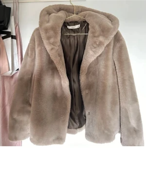 Beige pälsjacka med huva från Mango - Säljer en supermjuk beige pälsjacka från Mango med stor huva och diskreta knappar framtill. Jackan har en avslappnad passform och är helfodrad för extra komfort. Perfekt för dig som vill ha en cozy och trendig look under kyliga dagar.
