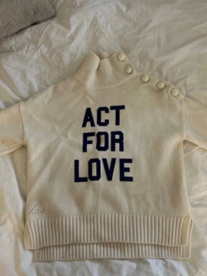 Krämvit stickad tröja Zadig & Voltaire - Säljer en krämvit stickad tröja från Zadig & Voltaire med hög krage och coola knappar längs axeln. Tröjan har texten 'ACT FOR LOVE' i mörkblått framtill och breda ribbade muddar. Perfekt för dig som gillar statement-plagg och snygga detaljer.  Lite små nopprig men syns på bild.