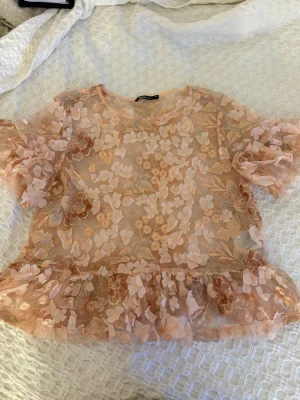 Blommig meshblus med volang - Supersöt blus i transparent mesh med 3D-blommor och broderade detaljer i beige och ljusrosa. Blusen har volangkant nedtill och puffiga korta ärmar. Blusen har används sparsamt och inga skador eller slitage. Storlek 36
