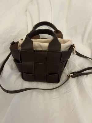 Brun flätad axelväska med canvaspåse - Säljer en snygg brun axelväska i flätad skinnimitation med avtagbar axelrem och handtag. Väskan har en innerpåse i beige canvas med dragsko. Perfekt storlek för det viktigaste och stilren design som sticker ut.