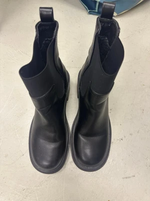Svarta chelsea boots från Super Me - Säljer ett par svarta chelsea boots från Super Me i storlek 37. Skorna har en rund tå, platt sula och är tillverkade i syntetmaterial med stretchiga partier på sidorna. Insidan är fodrad för extra värme och komfort. Perfekta till höst och vinter!