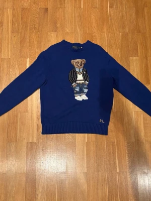Blå stickad tröja Polo Ralph Lauren - Blå stickad tröja från Polo Ralph Lauren med ikonisk björn på bröstet. Tröjan har rund hals, ribbade muddar och RL-broderi nertill. Perfekt för dig som vill ha en cool och klassisk look med en twist.