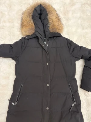 Svart lång pufferjacka Hollies - Säljer en svart lång pufferjacka från Hollies med stor huva och snygg äktapäls runt kanten. Jackan har dragkedja och knappar framtill, två fickor med dragkedja och quiltad design. Perfekt för kalla dagar och riktigt mysig att bära. Helt oanvänd. 