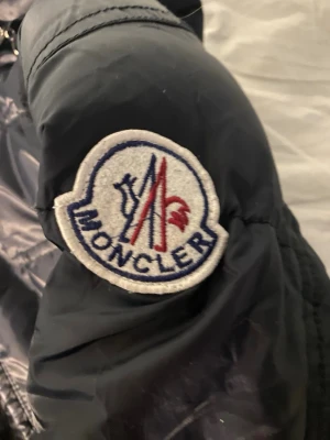 Svart Moncler lång pufferjacka - Säljer en svart lång pufferjacka från Moncler med huva och dragkedja. Jackan har quiltad design, två fickor med knappstängning och klassisk Moncler-logga på ärmen. Nypris: 9000