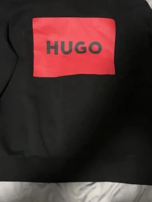 Svart sweatshirt från HUGO med röd logga - Svart sweatshirt från Hugo boss med stor röd logga på bröstet. Klassisk rund halsringning och ribbade muddar. Tröjan har en clean och modern look som passar perfekt till jeans eller joggers. Om du har några frågor är du alltid välkommen att skriva till mig.