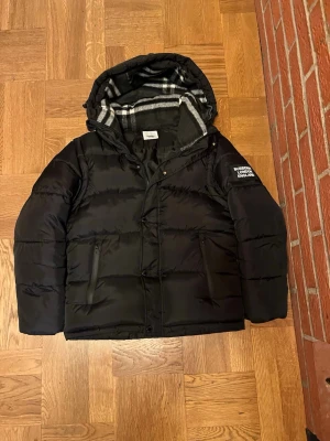 Svart pufferjacka från Burberry - Säljer en svart pufferjacka från Burberry med huva och klassiskt rutigt foder. Jackan har dragkedja, knappar och två fickor framtill. På vänster ärm finns en Burberry London England-logga. Perfekt för kalla dagar och riktigt snygg streetstil.