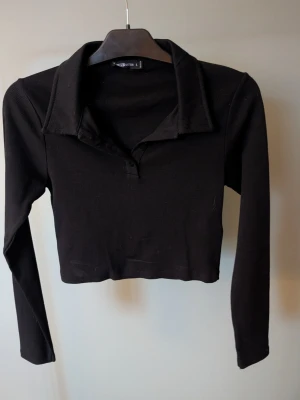Svart croppad långärmad topp - Svart croppad topp från FB. Sisters med lång ärm och klassisk krage. Toppen är ribbad och har knappar framtill vid halsen. Tillverkad i mjuk bomullsmix med lite stretch för en bekväm passform. Perfekt till jeans eller kjol för en cool look.