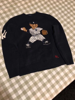 Mörkblå stickad tröja Ralph Lauren - Säljer en mörkblå stickad tröja från Ralph Lauren med broderad nalle i baseballkläder på bröstet och stort Yankees-märke på ärmen. RL-broderi i rött nertill. Perfekt för dig som gillar sportiga och unika plagg. Storleken är L men sitter som en S/M rekommenderar att man är 178-182 typ 65-75 kg. Jag är 184 väger 80 kg och den är lite för liten därför jag säljer den.