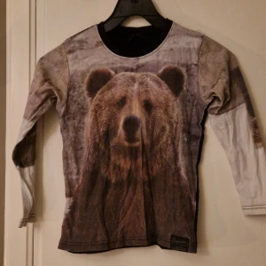 Långärmad t-shirt med björntryck - Unik långärmad t-shirt från Kids-Up med ett stort fototryck av en björn på framsidan. Tröjan har svarta detaljer vid halsen och ryggen, medan ärmarna är i beige och bruna toner. Tillverkad i mjuk bomull som känns skön mot huden. Storlek 116/122. Hund finns i hemmet. Kolla in mina andra annonser för samfrakt! 