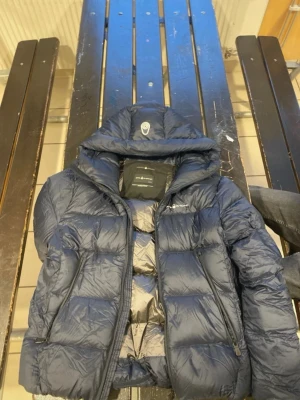 Mörkblå pufferjacka från Sail Racing - Säljer en mörkblå pufferjacka från Sail Racing i storlek medium. Jackan har huva, dragkedja framtill och två sidofickor med dragkedja. Perfekt för kalla dagar och riktigt skön att bära. Klassisk design med diskret logga på bröstet.