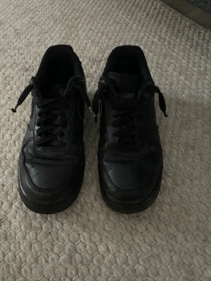 Svarta Nike Air Force 1 sneakers - Säljer ett par helsvarta Nike Air Force 1 sneakers i skinn med klassisk siluett och snörning. Skorna har rund tå, perforerade detaljer framtill och en platt sula. Perfekta för dig som gillar en clean och tidlös streetstil.