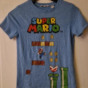 Blå Super Mario t-shirt från H&M - Säljer en blå t-shirt i storlek 134/140 från H&M med färgglatt Super Mario-tryck framtill. T-shirten har rund hals och korta ärmar, perfekt för dig som gillar gaming och retrostil. Trycket hade klistrat ihop sig lite längst ner vid tvätt, så det syns lite färgskiftningar vid de gröna rören (se bild). Hund finns i hemmet. Kolla in mina andra annonser för samfrakt! 