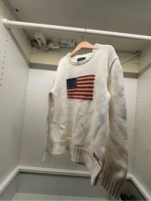 Vit stickad tröja Polo Ralph Lauren - Säljer en vit stickad tröja från Polo Ralph Lauren med amerikansk flagga broderad på bröstet och RL-broderi vid nederkanten. Tröjan har rund halsringning, ribbade muddar och lång ärm. Perfekt för dig som gillar klassisk preppy stil. Storlek S nytt pris 1399 