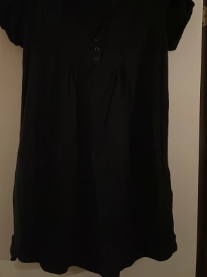 Svart kort klänning från Lindex - Säljer en svart kort klänning från Lindex med v-ringning och knappar framtill. Klänningen har korta ärmar och en enkel, rak passform. Perfekt till sommaren och lätt att styla med accessoarer. 100 procent bomull. Säljs på fler sidor.