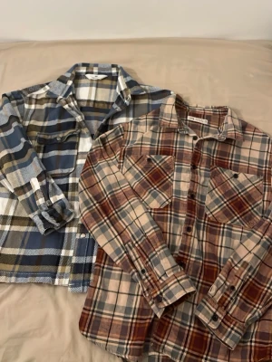 Två rutiga skjortor i bomull - Säljer två långärmade rutiga skjortor i mjuk bomull. Ena skjortan har blå, vit och olivgröna toner, medan den andra går i beige, brun och blått. Båda har klassisk krage, knappar framtill och bröstfickor. Perfekta för lager på lager och chill stil.