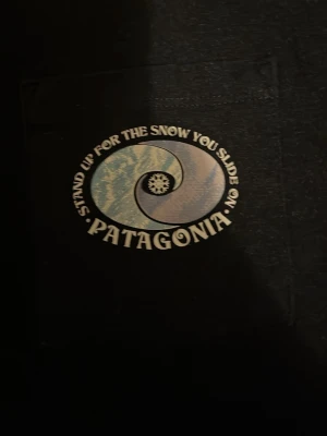 Svart Patagonia t-shirt med tryck XS - Svart t-shirt från Patagonia i regular fit med coolt tryck på bröstfickan och stor bergsgrafik på ryggen. Tillverkad av 50% återvunnen polyester och 50% återvunnen bomull. Perfekt för dig som gillar outdoor-vibbar och snygga prints.