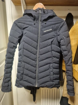 Svart skidjacka Peak Performance XS - Säljer en svart skidjacka från Peak Performance i storlek XS. Jackan har luva, dragkedja framtill och quiltade detaljer. Den har även snölås och fack för liftkort på armen.