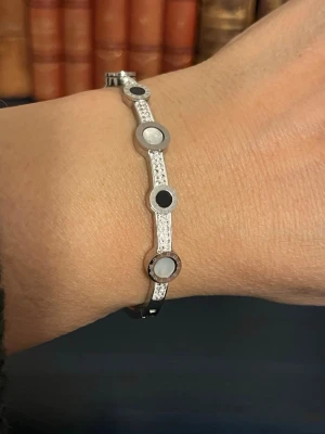 Silvrig armbågsformad bangle med stenar - Säljer ett stilrent och elegant silvrigt armband i bangle-modell med gnistrande stenar och svarta detaljer. Armbandet av rostfritt stål och har runda dekorationer med romerska siffror och pärlemorinslag. Perfekt accessoar för dig som gillar en clean men ändå lyxig look.
