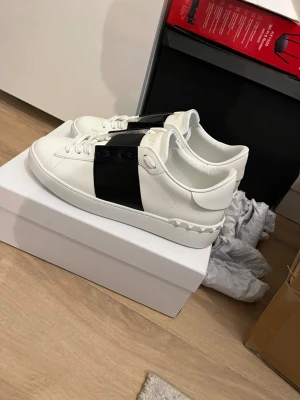 Valentino Garavani Open sneakers vit/svart - Säljer ett par Valentino Garavani Open sneakers i vitt skinn med bred svart läderdetalj över snörningen. Klassisk rund tå, vita snören och ikoniska nitar på hälen. Kommer med originalkartong och dustbag. Perfekta för dig som vill ha en clean men lyxig look.