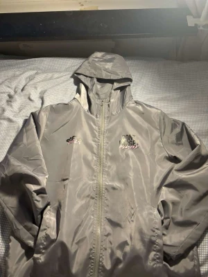 Grå vindjacka DFP Clothing M - Säljer en grå vindjacka från DFP Clothing i storlek M. Jackan har huva, hel dragkedja och tryck framtill med texten 'Don't Forget Palestine'. På ryggen finns ett stort rosa graffiti-inspirerat peace-tecken. Tillverkad i polyester och har en modern streetwear-vibe.