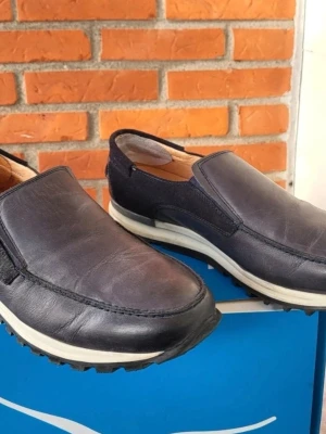 Mörkblå loafers från Cinnamon 44 - Säljer ett par mörkblå loafers från Cinnamon med ovandel i skinn och mocka. Skorna har en vit och svart sula med grovt mönster undertill och en klassisk slip-on design utan snörning. Insidan är fodrad i ljust läder för extra komfort.