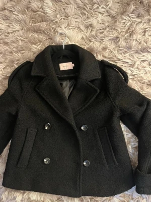 Svart teddy skepparkavaj från Nelly - Säljer en svart teddy skepparkavaj från Nelly med dubbelknäppning och stora kragar. Jackan har två fickor fram, axelklaffar och dekorativa knappar vid ärmsluten. Materialet är mjukt och fluffigt, perfekt för kyliga dagar. Helt ny! 