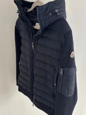 Moncler cardigan  - Säljer en svart pufferjacka från Moncler med stickade ärmar och huva. Jackan har dragkedja framtill, ribbade muddar och en ficka med dragkedja på ärmen. Perfekt för kyliga dagar och riktigt snygg streetstil.