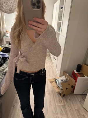 Beige omlott kofta med knyt - Supersnygg beige omlott kofta med knyt i sidan och v-ringad hals. Tröjan är stickad och har långa ärmar samt croppad passform. Perfekt till jeans eller kjol för en chill men ändå stilsäker look.