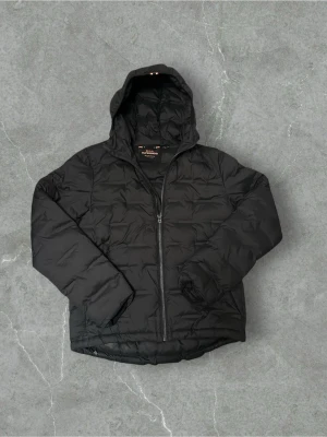 Svart pufferjacka från Superdry - Säljer en svart pufferjacka från Superdry Studios med huva och dragkedja framtill. Jackan har en clean look utan synliga loggor på framsidan, men en diskret patch på ärmen. Perfekt för kalla dagar och riktigt skön att bära.