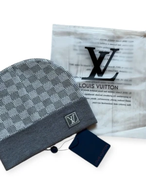 Grå rutig mössa från Iv - Snygg grå mössa från Iv med klassiskt rutigt mönster och vikt kant. Mössan har en diskret Iv-logga framtill och är tillverkad i mjukt material som känns lyxigt. Perfekt accessoar för att lyfta din vinterstil.