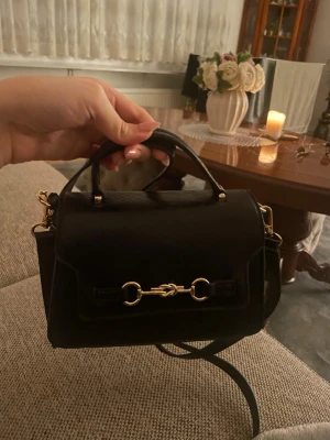 Svart handväska med gulddetaljer - Snygg svart handväska i skinnimitation med struktur och guldfärgade metalldetaljer. Väskan har både handtag och axelrem, samt ett dekorativt spänne framtill. Perfekt storlek för det viktigaste och stängs med magnetlås. Den är alltså även helt ny och oanvänd!!