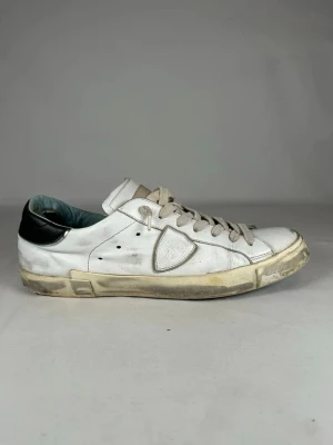 Vita sneakers från Philippe Model - Säljer ett par vita sneakers från Philippe Model med svart detalj på hälen och grå snörning. Skorna har en klassisk låg profil, rund tå och är tillverkade i skinn. Sidan pryds av märkets ikoniska sköldlogga. Perfekta för dig som gillar stilrena och tidlösa sneakers.