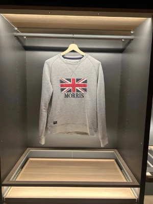 Grå sweatshirt från Morris med flagga - Säljer en grå sweatshirt från Morris i bomull med Union Jack-flagga och broderad logga på bröstet. Tröjan har rund halsringning, långa ärmar och klassisk passform. Perfekt för dig som gillar stilren och brittisk-inspirerad design.(den har en liten defekt vid den ena axeln se sista bilden).        Längd:66cm.     Bredd:46cm