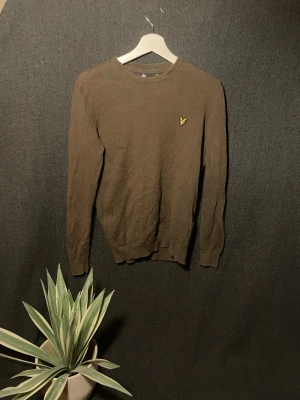 Olivgrön stickad tröja Lyle & Scott - Snygg olivgrön stickad tröja från Lyle & Scott i storlek S. Klassisk rund hals och långärmad modell med den ikoniska gula fågeln broderad på bröstet. Perfekt för dig som gillar stilrena och tidlösa plagg med en modern touch.