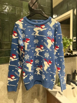 Blå jultröja Pull&Bear med skidåkare - Blå stickad tröja från Pull&Bear med vita skidåkare i röd tomteluva och snöflingor över hela tröjan. Rund halsringning och ribbade muddar. Perfekt för dig som vill sticka ut på julmiddagen eller vintermyset. Pris är diskuterbart vid en snabb affär!