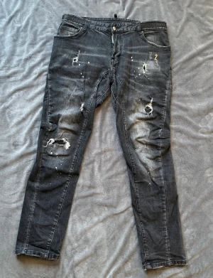 Svarta Dsquared2 jeans med slitningar - Säljer ett par svarta jeans från Dsquared2, modell Classic Kenny Twist Jean. De har snygga slitningar, färgstänk och en snygg tvättad look. 